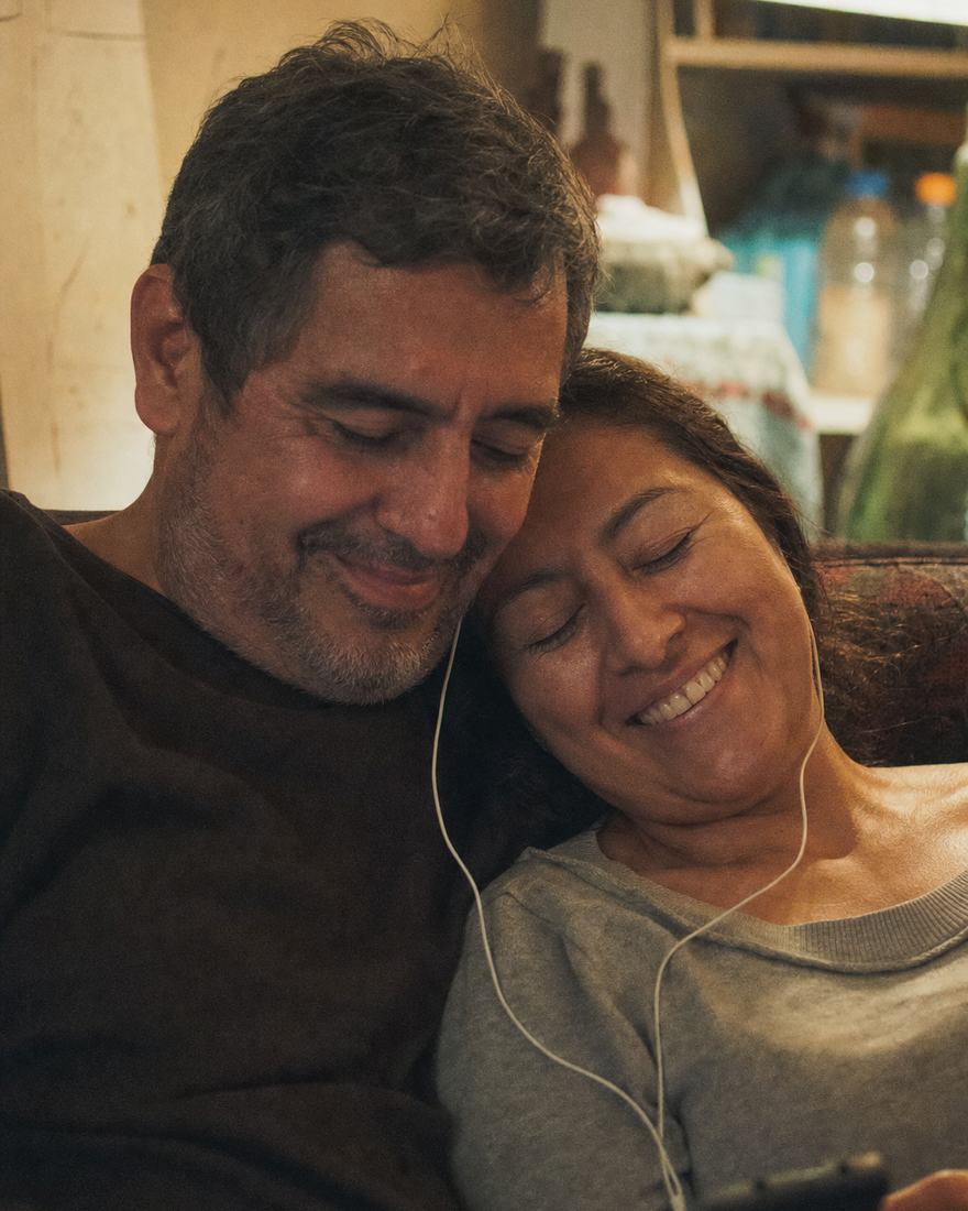 Una pareja de casados de muchos años comparte unos audífonos en el sofá, ella recostada en él con los ojos cerrados